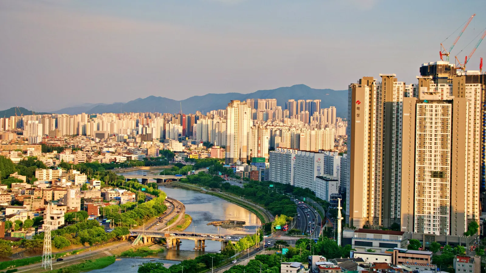 Aerial view of Daegu's high-rises along a river at golden hour, mountains on the horizon
