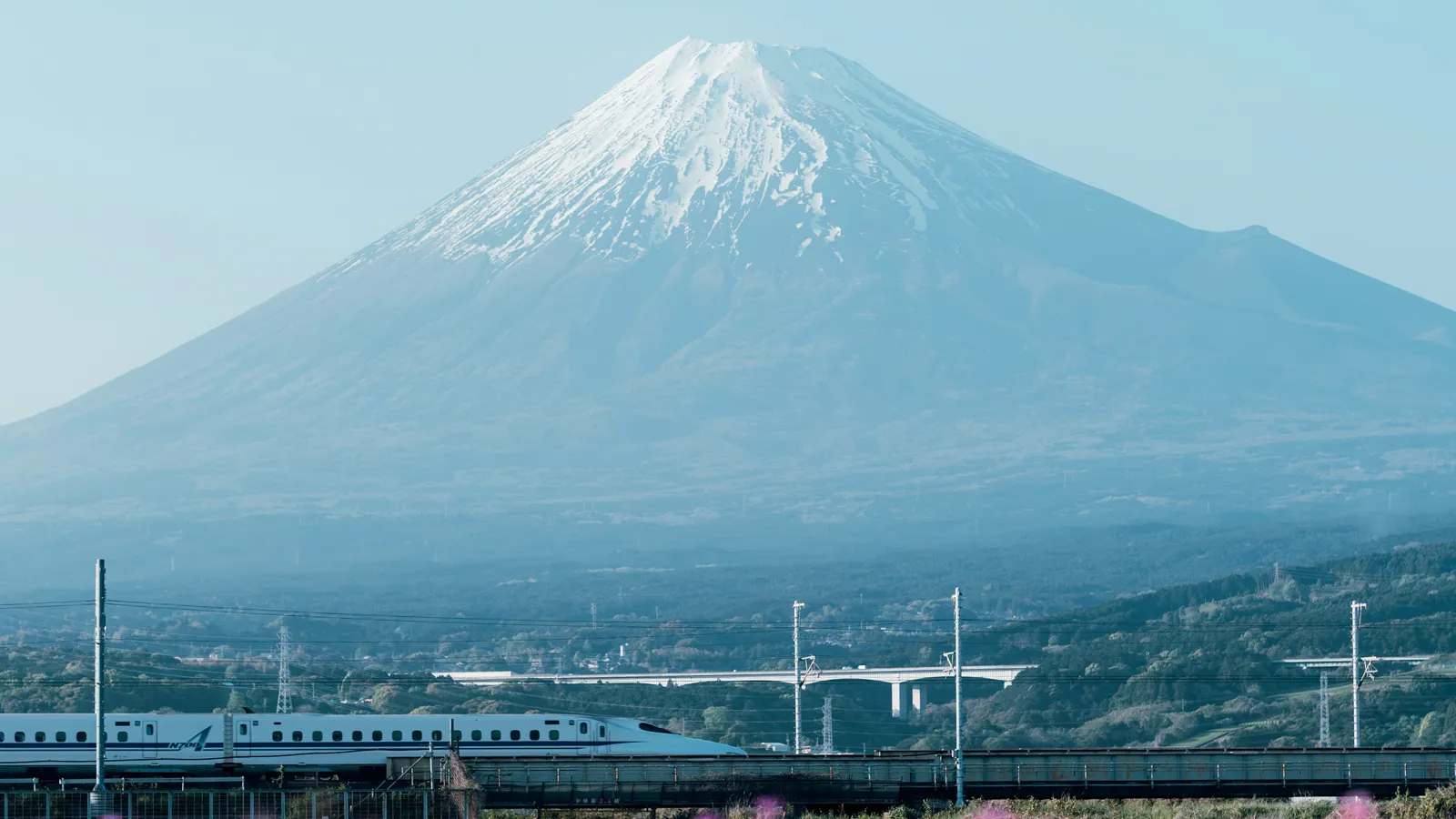 A Shinkansen train crossing a viaduct with snow-capped Mount Fuji rising behind it